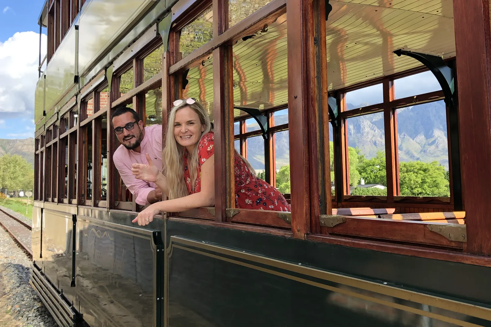 Two travellers leaning from the window of a vintage tram, vines and mountains behind